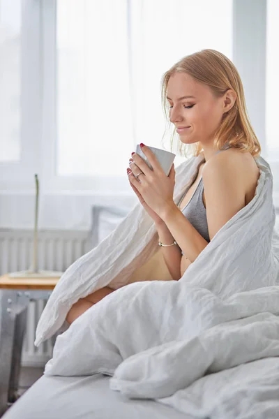 Pretty young woman with cup of tea in bed — Stockfoto