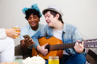 Multiethnic university roommates singing and reading lyrics in cell phone screen in small apartment at living room with food snacks and drinks. Carefree, happiness and bonding feelings clipart