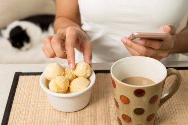 Close up of woman picking balls of bread cheese (known as Pao de Queijo in Brazil) and holding cell phone. Cup of coffee and comestic cat in the background. clipart