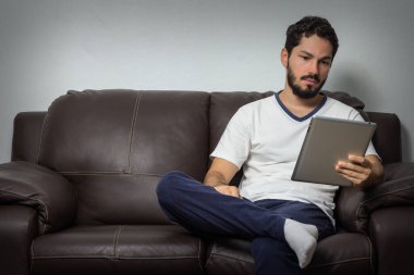 Evdeki kanepede elinde tabletle oturan adam.