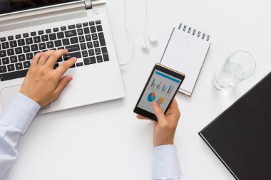 Genç yönetici aynı anda hem cep telefonu bilgilerine göz atıyor hem de dizüstü bilgisayarda yazıyor. Kişinin bakış açısı. Beyaz tablo.