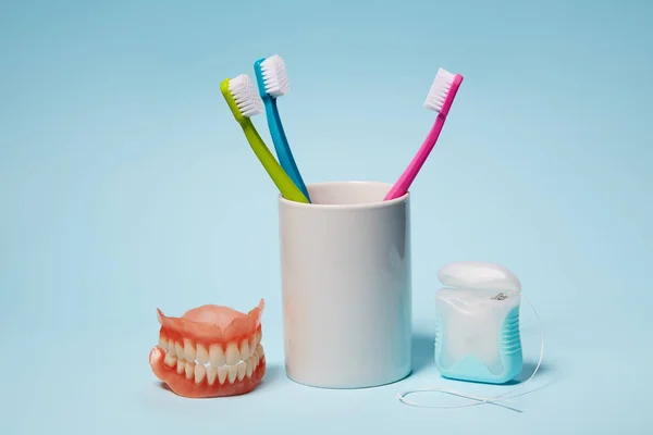 Colorful toothbrushes, dentures and dental floss on light blue background