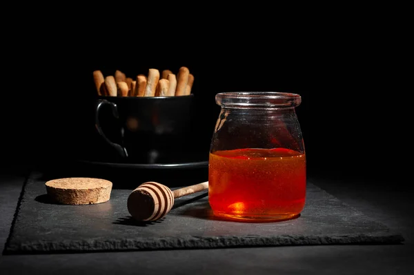 Jar with fresh honey and a wooden spoon for honey with bread crumbs for dessert on a serving black board on a black background — Stock Photo, Image