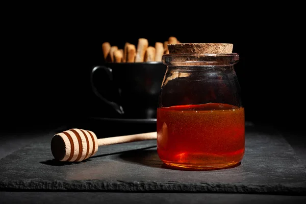 Jar with fresh honey and a wooden spoon for honey with bread crumbs for dessert on a serving black board on a black background — Stock Photo, Image