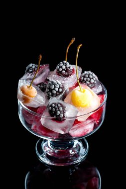  Donmuş ahududu kirazları ve böğürtlenler siyah arka planda cam bir kasede buz parçalarındalar.