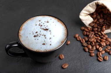  Beyaz köpüklü siyah bir fincan kahve, dağıtım için siyah bir tahta üzerinde duruyor, kahve çekirdekleri orada dağılıyor. kopyalama alanı