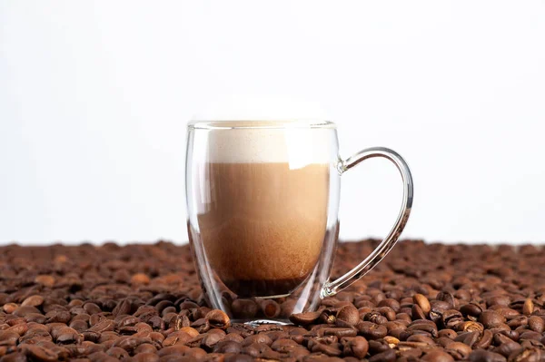 Uma xícara de latte transparente de fundo duplo fica entre os grãos de café em um fundo branco. Espaço de cópia — Fotografia de Stock