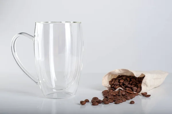 Empty transparent mug with a double bottom, next to which coffee beans spilled out of a bag on a white background. Copyspace — Stock Photo, Image