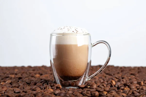 Uma xícara transparente de latte com canela em uma espuma com um fundo duplo fica entre os grãos de café em um fundo branco. Espaço de cópia — Fotografia de Stock