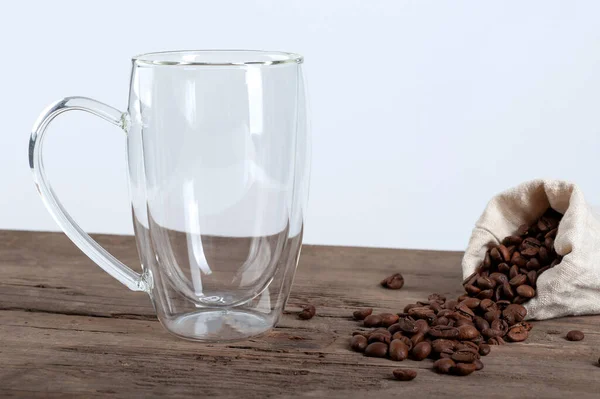 An empty transparent mug with a double bottom stands on an old wooden background, next to which coffee beans against a white background. copyspace — Stock Photo, Image