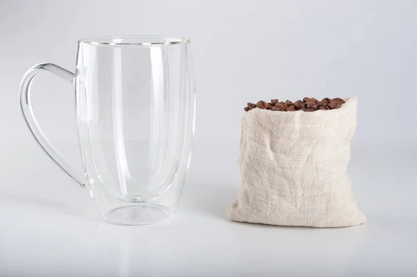 Empty transparent mug with a double bottom, next to which coffee beans against a white background. Copyspace — Stock Photo, Image