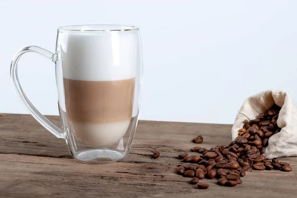 Latte em uma caneca transparente fica em uma mesa de madeira em um fundo branco, ao lado de grãos de café. espaço de cópia — Fotografia de Stock