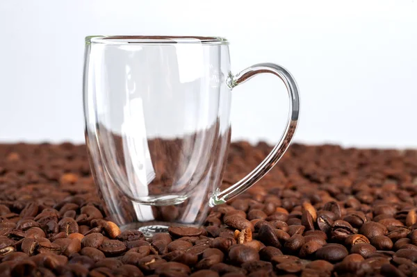 Une tasse transparente vide à double fond se dresse parmi les grains de café sur un fond blanc. Espace de copie — Photo