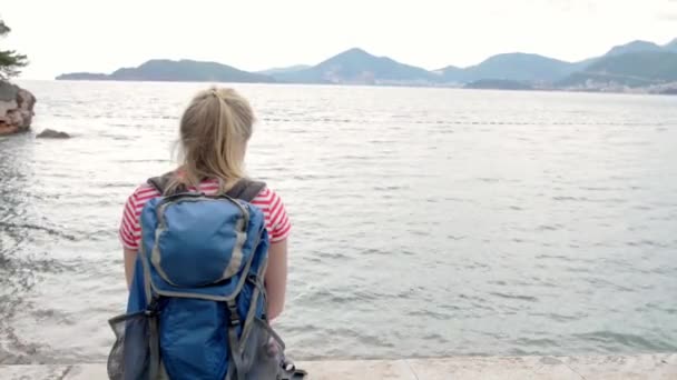 Młoda blondynka z workiem patrzy na morze — Wideo stockowe