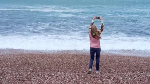 Молодая девушка тысячелетие дробит море в облачную погоду на своем смартфоне — стоковое видео