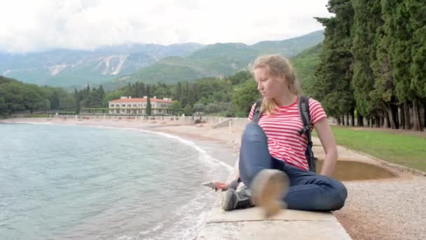 Κορίτσι κάθεται και σαρώνει στο smartphone κοντά στη θάλασσα και τα βουνά — Αρχείο Βίντεο