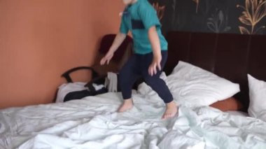 Beyaz çocuk 4 yaşında, büyük beyaz bir çift kişilik yatağın üstünde zıplıyor.