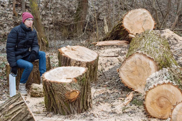 Hard work done: young woman saws a huge tree in the forest and sits resting with a saw in her hand