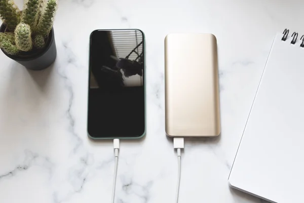Portatif bir güç bankasından kaktüs ve not defteriyle mermer bir masa zemininde şarj olan düz bir akıllı telefon.