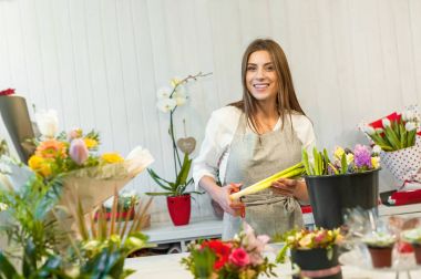 Smiling woman florist small business flower shop owner, at counter holding a scissor cuting hyacinths, making arrangements and smiling at camera. clipart