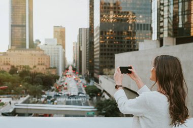 Güzel genç kadın bir köprüde kentsel sahne, şehir merkezinde, boulevard boyunca gün batımında, alarak fotoğraf smartphone ile oturuyordu.