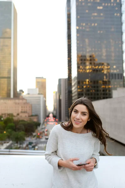 Beautiful young woman sitting on a bridge across the boulevard in urban scenery, downtown, at sunset, taking photos with smartphone and smiling at camera.