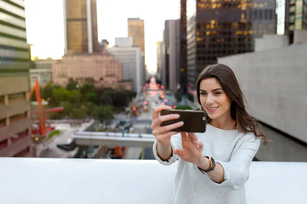 美丽的年轻女子坐在横跨林荫大道的桥上 在市中心 日落时分 用智能手机拍摄自画像 — 图库照片