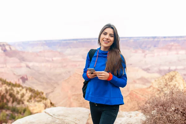 Travel hiking photo of young beautiful woman with backpack at Grand Canyon viewpoint, Arizona, USA smiling at camera and holding a smartphone.