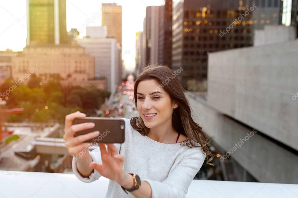 Beautiful young woman sitting on a bridge across the boulevard in urban scenery, downtown, at sunset, taking self portraits with smartphone.