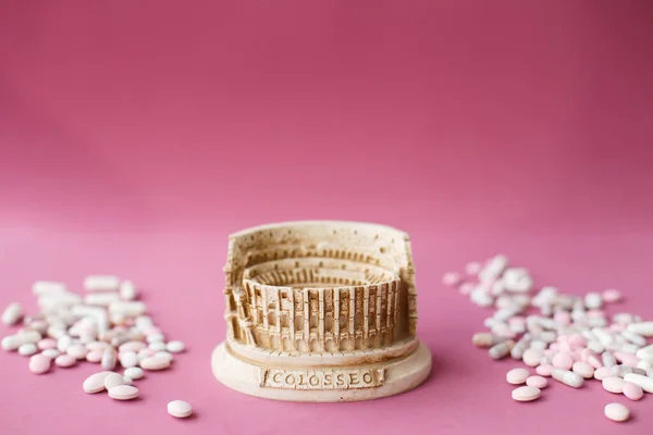 Stone statuette of the Coliseum building in Rome in Italy, surrounded by a bunch of antiviral pills and tables. Concept of dangerous infection with the Chinese coronavirus covid-2019. Pink background.