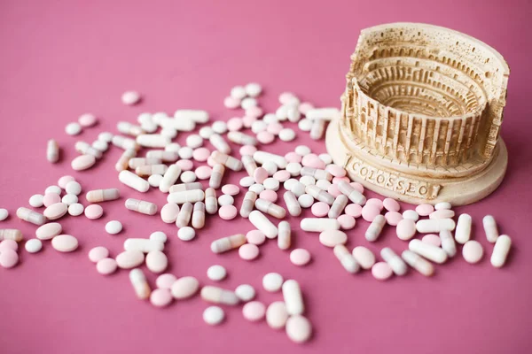 Stone statuette of the Coliseum building in Rome in Italy, surrounded by a bunch of antiviral pills and tables. Concept of dangerous infection with the Chinese coronavirus covid-2019. Pink background.
