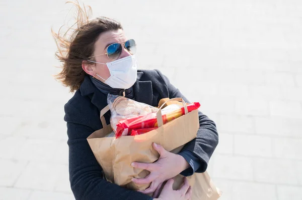 Scared woman in protective face mask,gray coat.Making store of food in craft bag before isolation:toilet paper,paste,cookies.Lack of products.Leaving city during quarantine of coronavirus covid-2019.