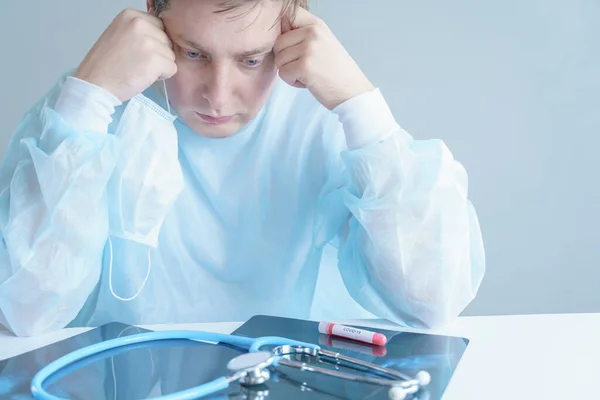 Sad male doctor in face mask, medical robe, tired from work. In horror,despair from treating sick patients.Stethoscope, blood test tube.Fighting coronavirus covid-19.Dangerous infection. Stay at home.