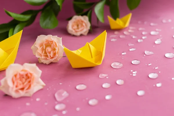 Yellow spring paper ships,boats floating in sparkling water drops on purple background.Pink full roses,branch of green leaves.Summer concept.Greeting card.DIY family activity entertainment with kids.