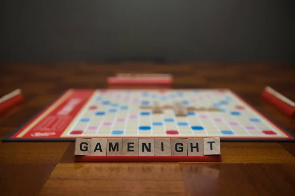 Brev kakel stavning ord spel natt på stativ i förgrunden med ur fokus spelplan i bakgrunden. — Stockfoto