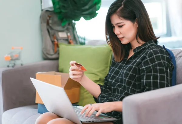 Asyalı genç bir kadın kredi kartı tutuyor ve evde oturma odasında online alışveriş yapmak için dizüstü bilgisayar kullanıyor.