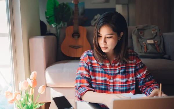 Beautiful asian young woman working from home on laptop computer while sitting at condo living room with confidence posing in coronavirus or covid-19 outbreak situation