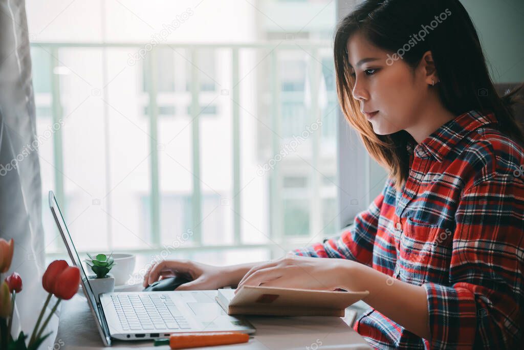 Beautiful asian young woman working from home on laptop computer while sitting at condo living room with confidence posing in coronavirus or covid-19 outbreak situation