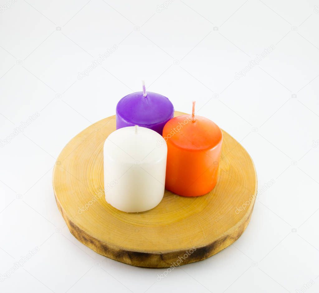 Three candles (white, lilac, orange) stand on a wooden stand from a cut of a tree trunk. White background.