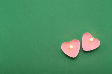 Yeşil arka planda kalp şeklinde yanan iki pembe mum. Metal bir kapta mumlar.