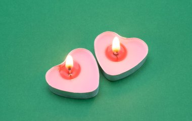 Yeşil arka planda kalp şeklinde yanan iki pembe mum. Metal bir kapta mumlar.