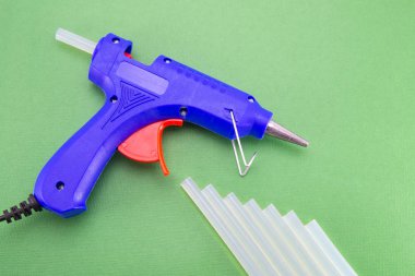 Yeşil arka planda sıcak erime yapıştırıcısı için yeni mavi yapıştırıcı tabancası. (masa). Şeffaf renk tabancasının sapları yakınlarda duruyor. El sanatları ve tamirat için yüksek kaliteli bir araç. Elektrikli el aracı.