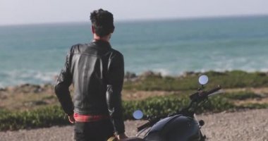 Yakışıklı motorcu klasik motosikletten deniz kenarına doğru yürüyor.