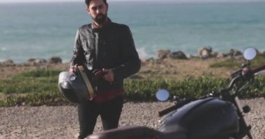 Deniz kenarından yürüyen yakışıklı motorcu klasik motorsiklete biniyor.