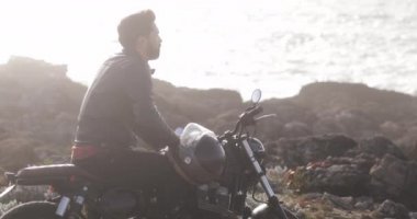 Yakışıklı bir adam klasik bir motosiklette oturmuş, deniz kenarına bakıyor.