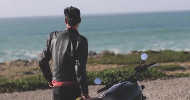 Handsome biker man walking from classic motorcycle towards the seafront — Stock Video