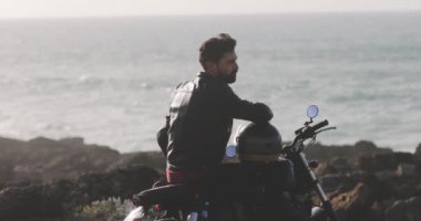 Yakışıklı adam deniz kenarında klasik bir motosiklette oturuyor.