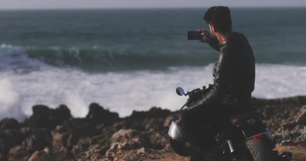 Snygg man som sitter på motorcykel och fotograferar havet — Stockvideo