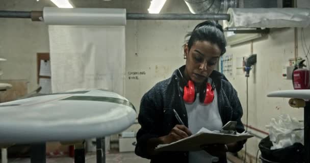 Mujer negra escribiendo en portapapeles chequeando tabla de surf en taller — Vídeos de Stock