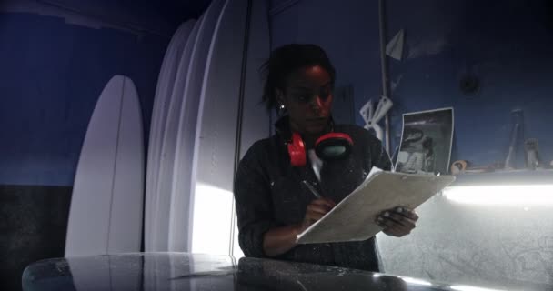 작업실에서 클립보드에 글씨를 쓰고 파도타기 판을 확인하는 흑인 여자 — 비디오
